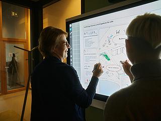 Innenaufnahme zeigt die Silhouette zweier Frauen, die an einem Smartboard mit dem Rücken zur Kamera stehen und mit Stiften ein Bild markieren.