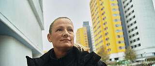 Außenaufnahme zeigt eine Hausmeisterin der GESOBAU unterwegs im Märkischen Viertel. Rechts hinter der Frau sind unscharf zwei markante, gelb getünchte Hochhäuser im Märkischen Viertel erkennbar.