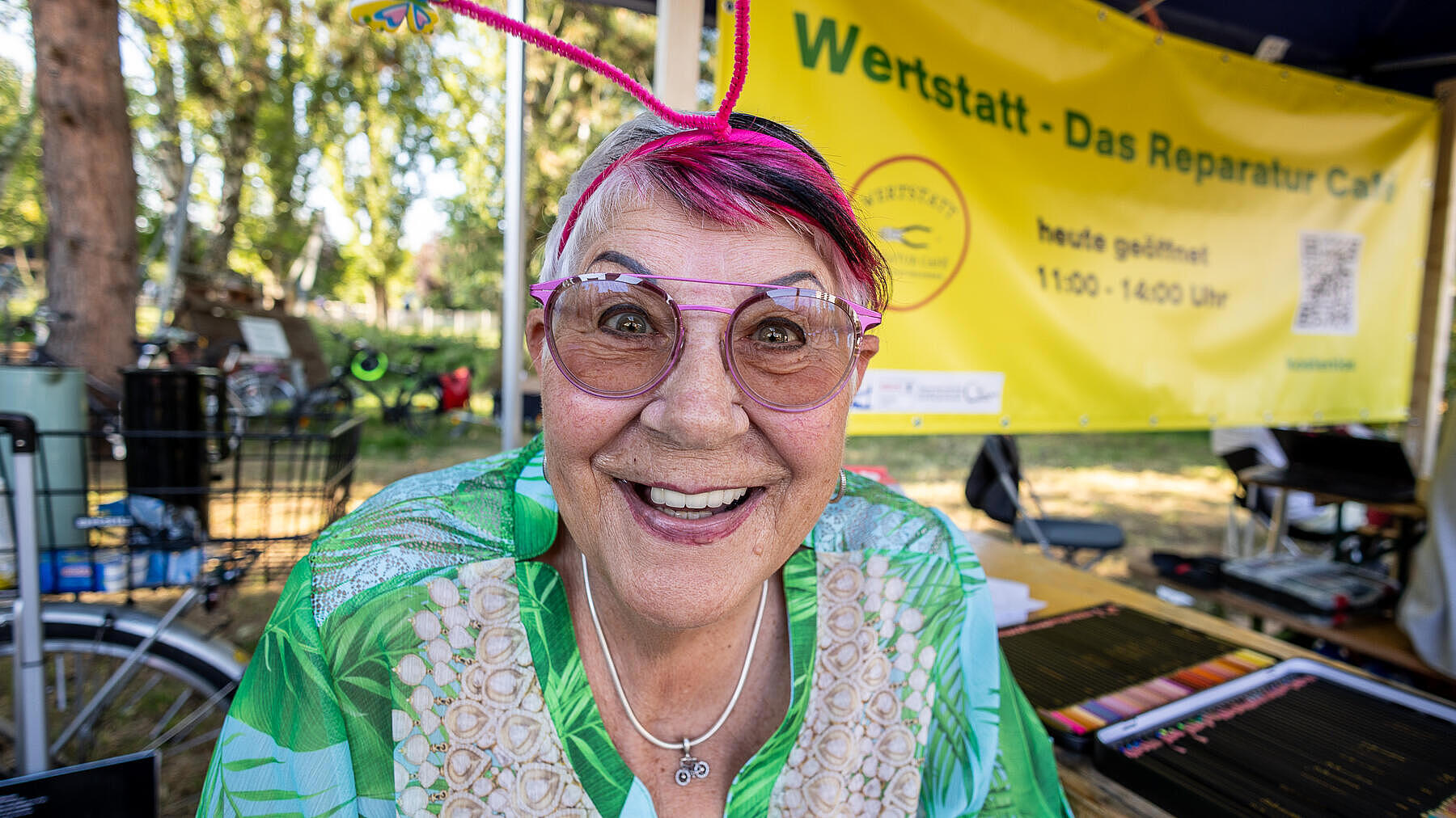 Außenaufnahme zeigt eine in die Kamera lachende, ältere Frau mit pinker Brille, pinken Haaren und einem selbstgebastelten Haarschmuck. 