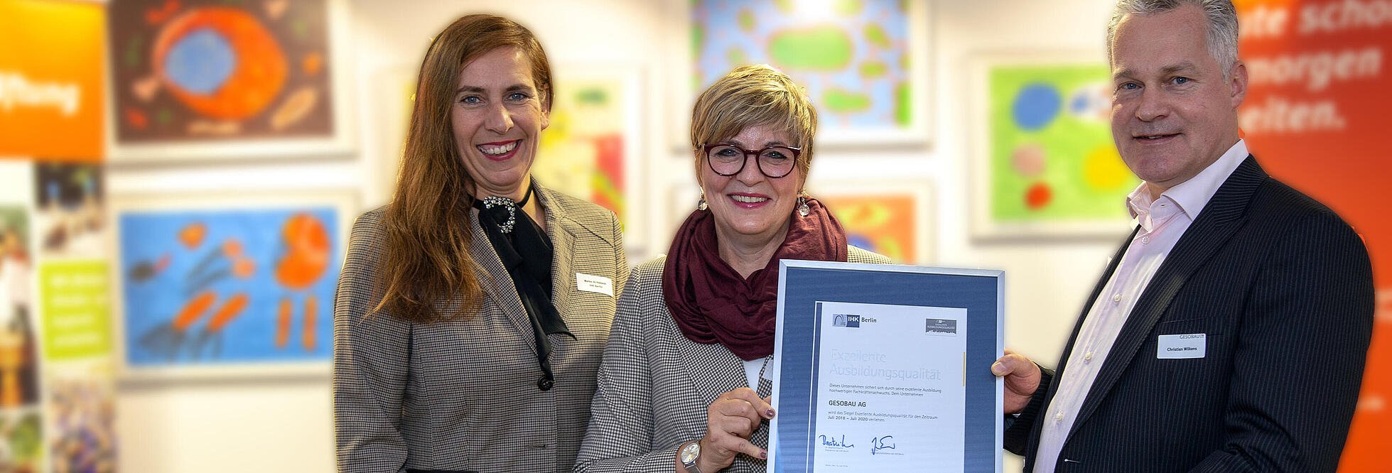 Innenaufnahme zeigt eine weiblich gelesene Person der IHK sowie Petra Ebert und Christian Wilkens mit einer Urkunde der IHK Berlin