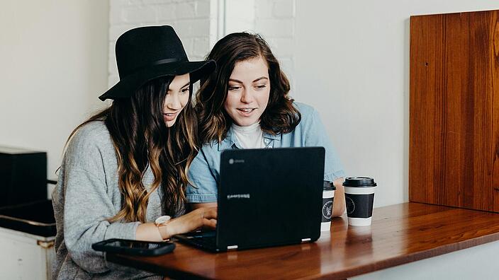 Zwei junge Frauen sitzen vor einem augeklappten Laptop an einem Holztisch.