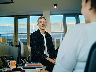 Innenaufnahme zeigt zwei Kollegen in einem Gespräch. rechts mit dem Rücken zur Betrahcterin sieht man die Schulter und Teile des Profils einer lächelnden Kollegen, mittig ein junger Mann mit Brille, der auf einem Schreibtisch sitzt und lächelt.
