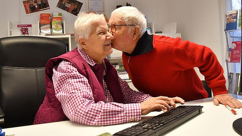 Senior küsst Seniorin auf die Wange, beide sitzen am PC
