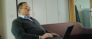 Innenaufnahme zeigt einen GEOSBAU-Mitarbeiter auf einem Sofa sitzend und leicht nach hinten gelehnt, auf seinem Schoß ein aufgeklappter Laptop, an dem dieser konzentriert arbeitet.