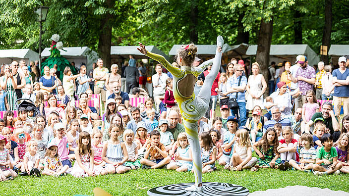Außenaufnahme zeigt eine im Halbkreis stehende und teilweise sitzende Menschenmenge auf der Wiese im Schloßpark Schönhausen von der Perspektive des Zirkus' gesehen. Im Hintergrund einige Stände des Kunstfestes. 