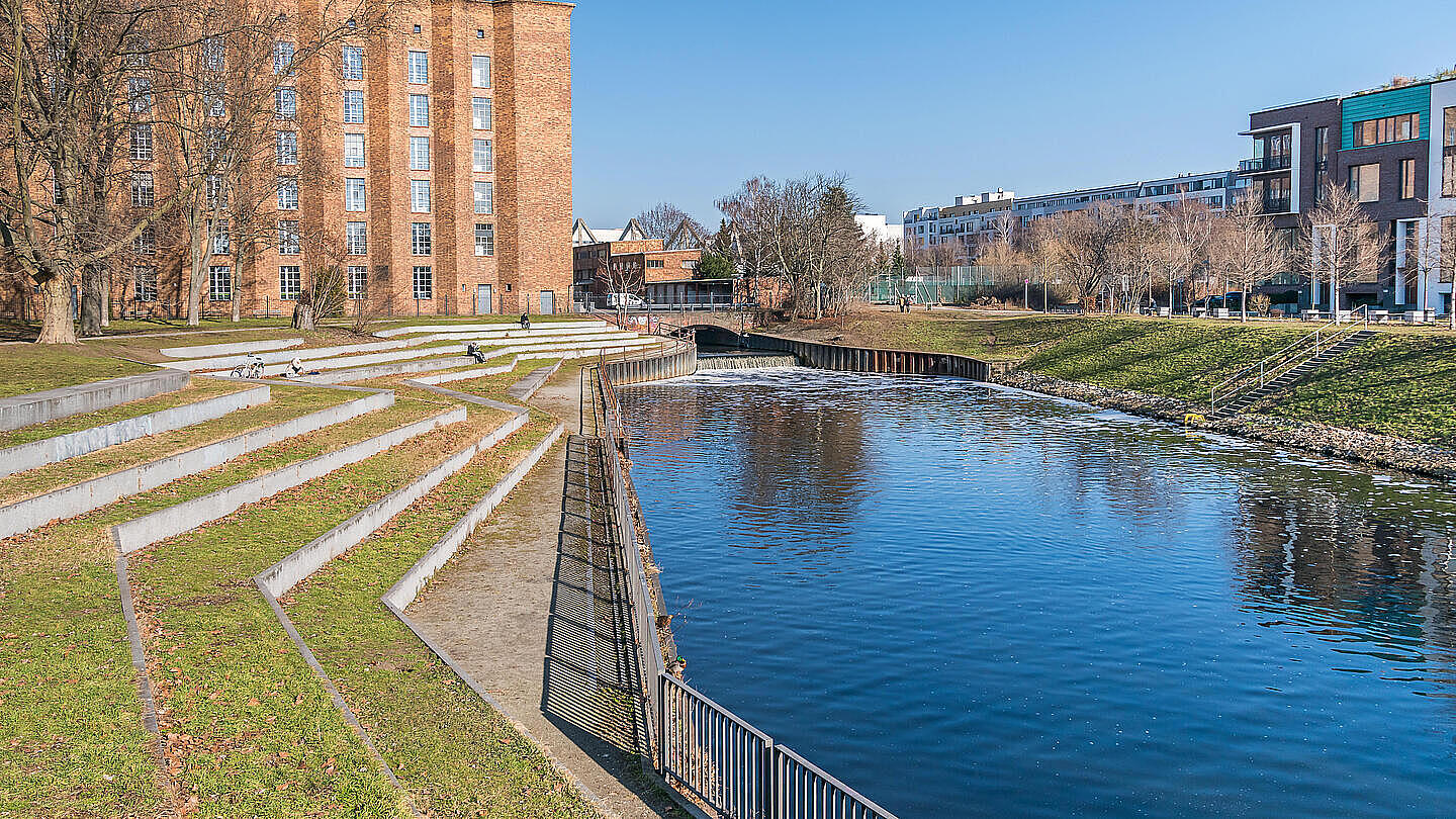Die Außenaufnahme zeigt einen Ausschnitt des Sellerparks in Berlin-Mitte. Die Seiten des Kanals, der sich durch das Bild zieht, ist gesäumt von Rasen und Bäumen. Im Hintergrund links befindet sich ein großes Backsteingebäude und auf der rechten Seite Wohnhäuser.
