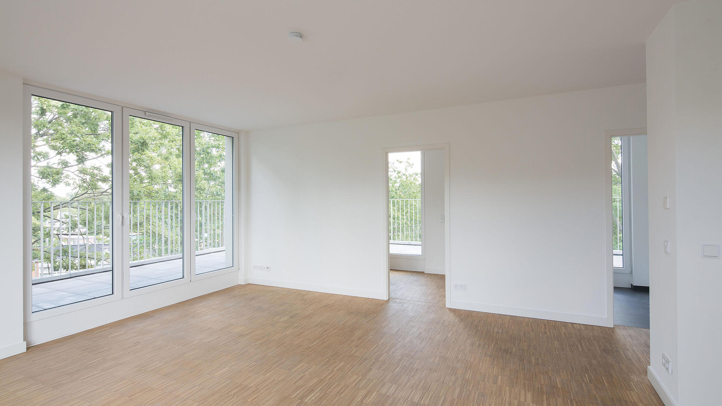 Innenaufnahme zeigt ein leeres Zimmer mit hellem Stabholzparkett, bodentiefen Fenstern und umlaufendem Balkon.