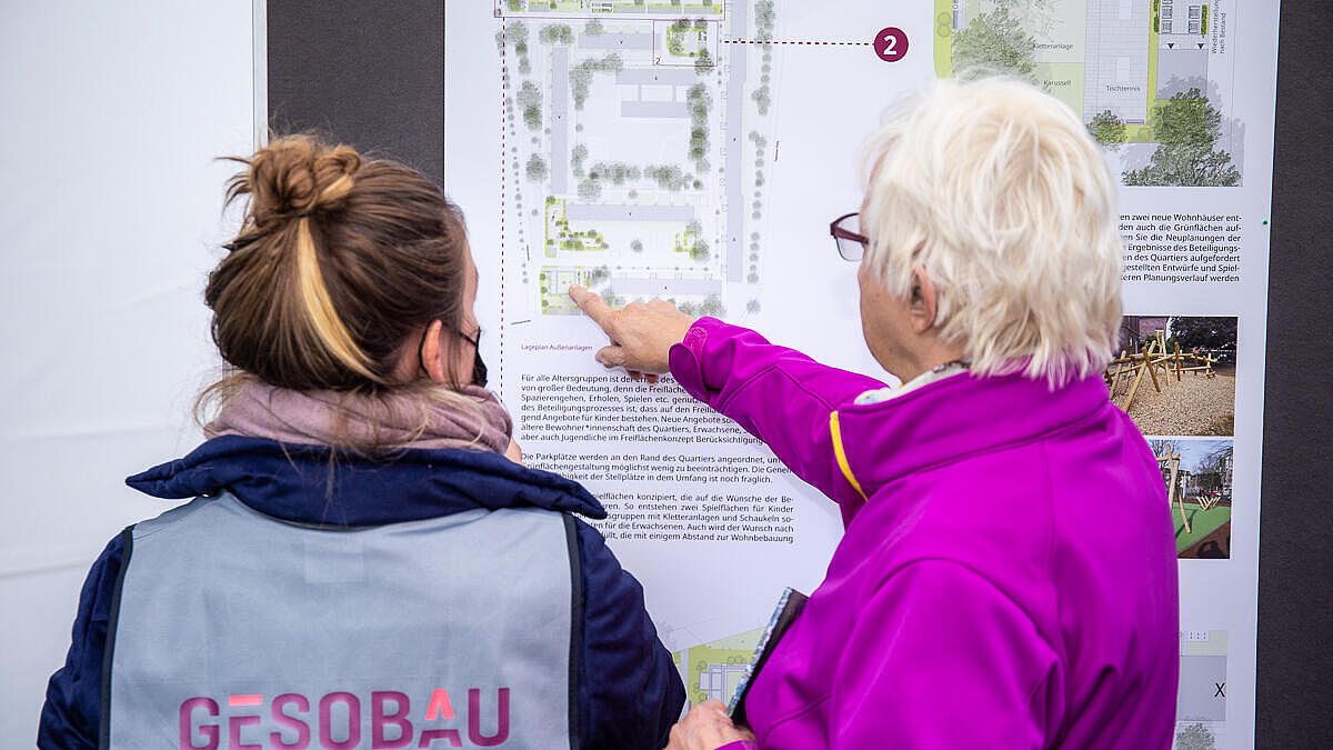 Zwei Personen stehen einem Informationsplakat zugewandt, die links stehende trägt erkennbar eine Weste mit dem GESOBAU-Logo, die rechte deutet mit ihrem rechten ausgetreckten Zeigefinger auf das Plakat.