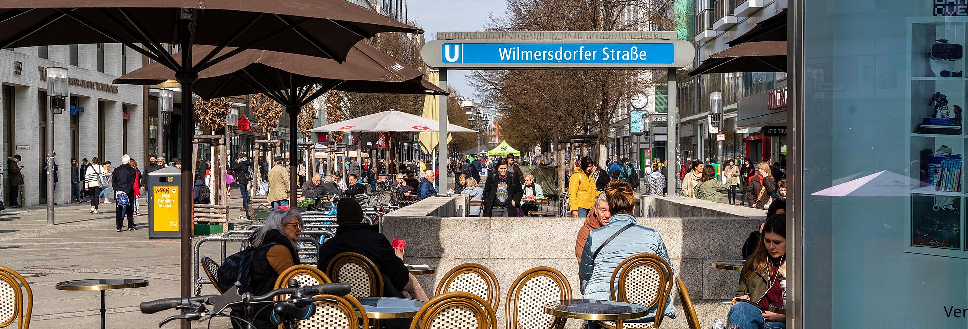 Die Außenaufnahme zeigt die Wilmersdorfer Straße im Bezirk Berlin-Wilmersdorf. Zu sehen ist eine belebte Straße mit bummelnden Menschen und sich aneinaderreihenden Cafés. Manche Personen nehmen den Abgang in die U-Bahn-Station.