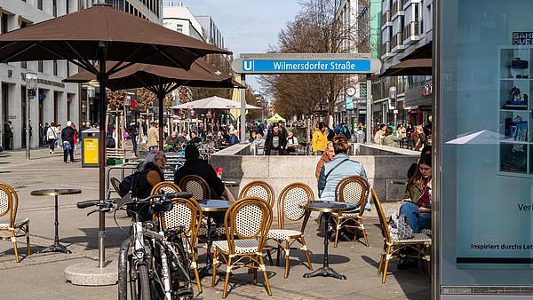 Die Außenaufnahme zeigt die Wilmersdorfer Straße im Bezirk Berlin-Wilmersdorf. Zu sehen ist eine belebte Straße mit bummelnden Menschen und sich aneinaderreihenden Cafés. Manche Personen nehmen den Abgang in die U-Bahn-Station.