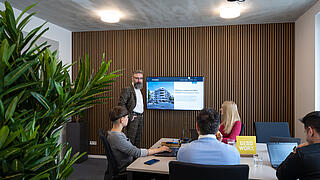 Innenaufnahme zeigt einen Meetingraum in der Langhansstraße 28 mit drei um einen Tisch sitzenden und einer an einem Whiteboard stehenden Person.