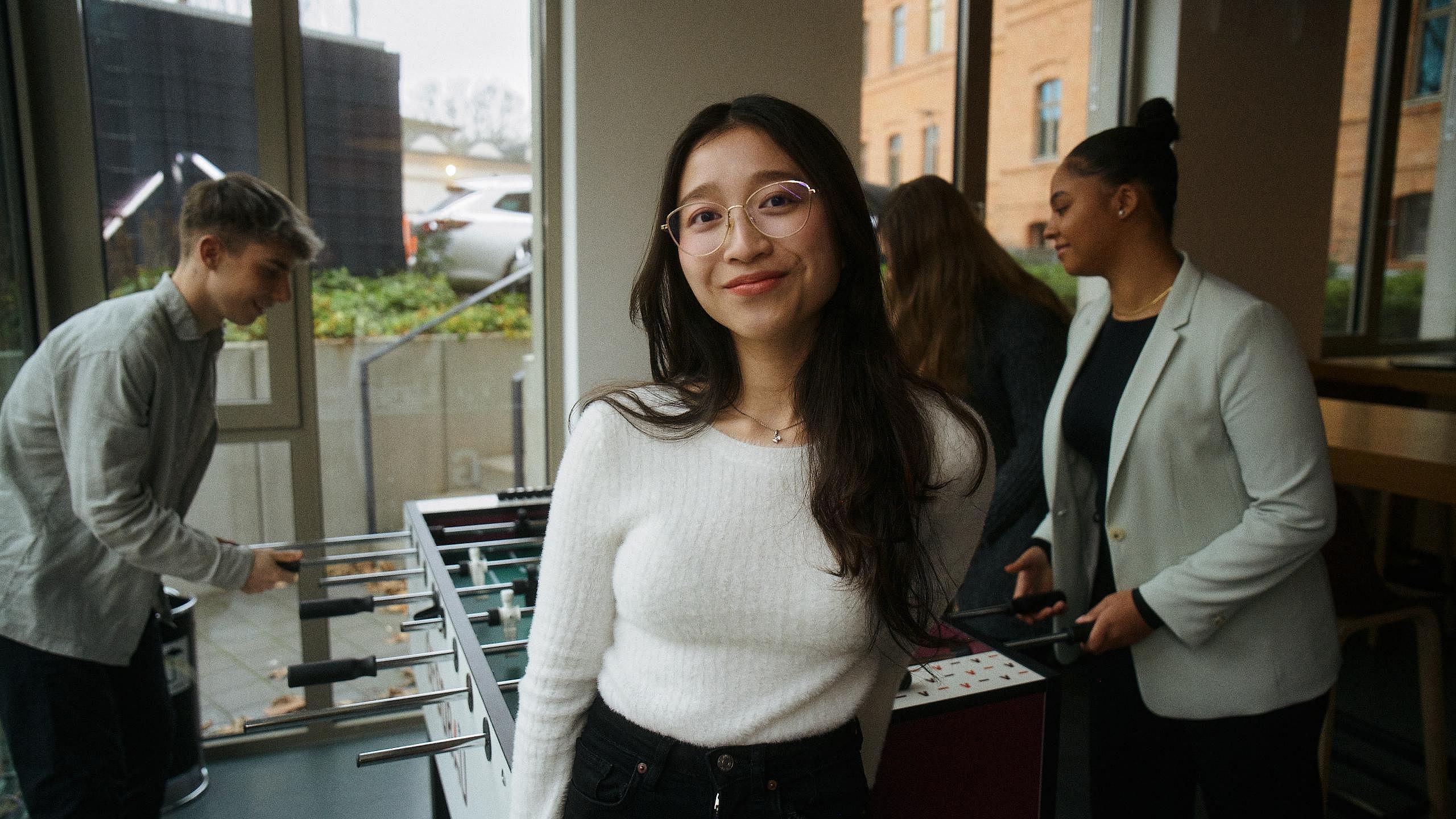 Innenaufnahme zeigt eine Gruppe von vier Auszubildenden, die an einem Tischkicker spielen. Mittig steht eine der vier mit dem Rücken zum Kicker und lächelt selbstbwusst in die Kamera.