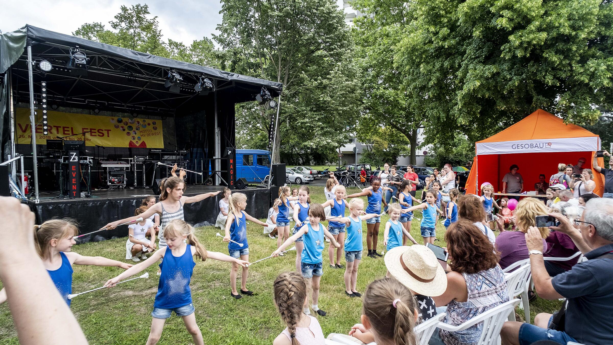 Außenaufnahme zeigt eine Gruppe von in Formation tunrnenden Kindern eines lokalen Sportvereins vor der Bühne des VIERTEL FEST bei sommerlichen Temperaturen. Rechts in der Ecke sitzend das Publikum, im Hintergrund ein Pavillon der GESOBAU.
