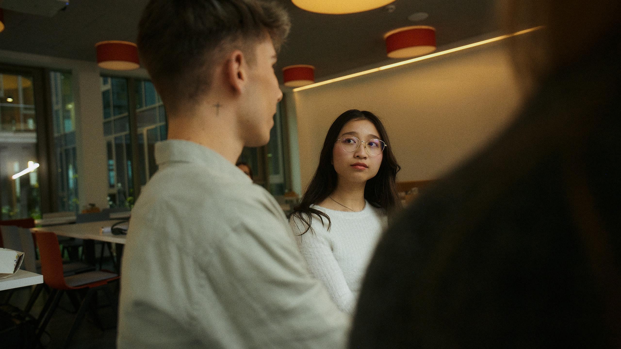 Innenaufnahme zeigt mittig eine sehr junge Auszubildende mit Brille, die konzentriert einem uns nur im Profil sichtbaren, männlichem Azubi zuhört. Dieser hat hinter seinem Ohr ein kleines, leicht sichtbares Tattoo. Beide Personen sind in einer Kantine.