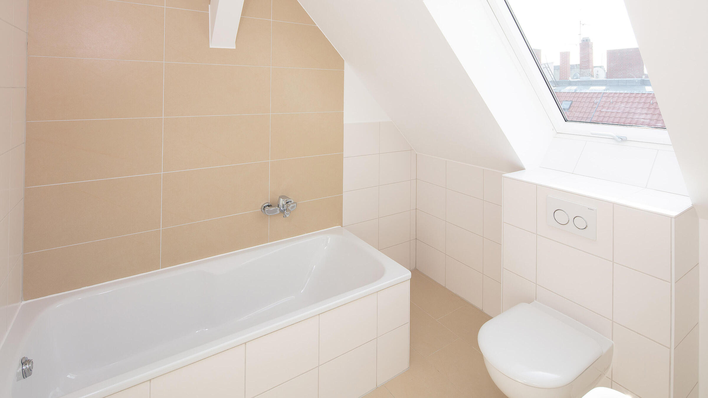 Innenaufnahme zeigt ein typisches Bad der neu gebauten Dachgesosswohnungen: linker Hand eine Badewanne, rechts unter einem schrägen Dachfenster die Toilette mit Spülkasten, in der vorderen Ecke des Fotos ist eine Ecke des Handwaschbeckens sichtbar.