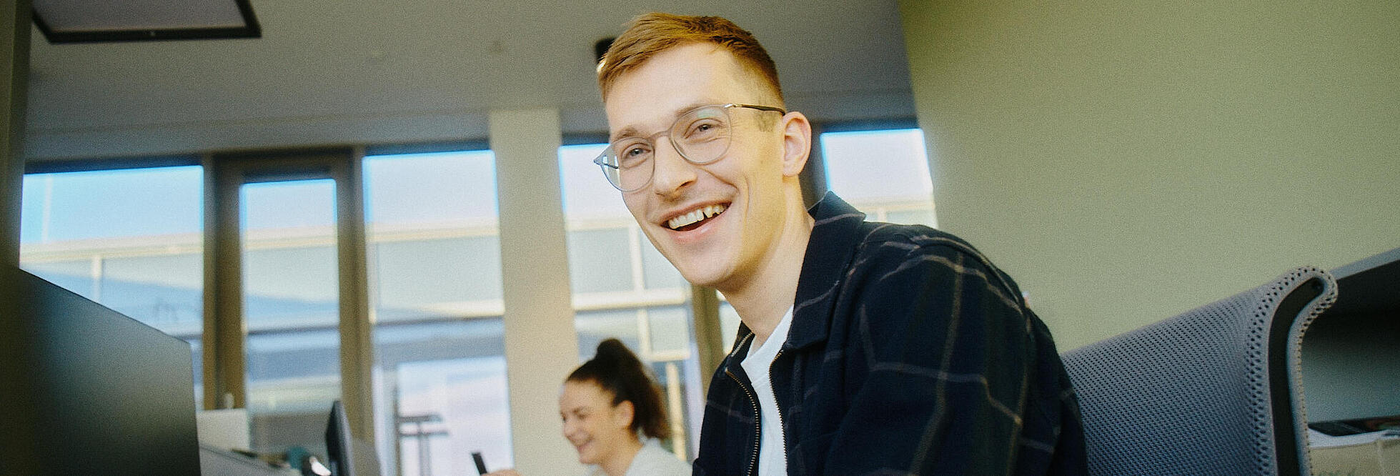 Innenaufnahme zeigt Mitarbeitende der GESOBAU an ihren Bürotischen sitzend. Im Vordergrund ein Kollege, der lächelnd in die Kamera blickt. Im Hintergrund eine lächelnde Kollegin, die etwas in ihr Smartphone eingibt.
