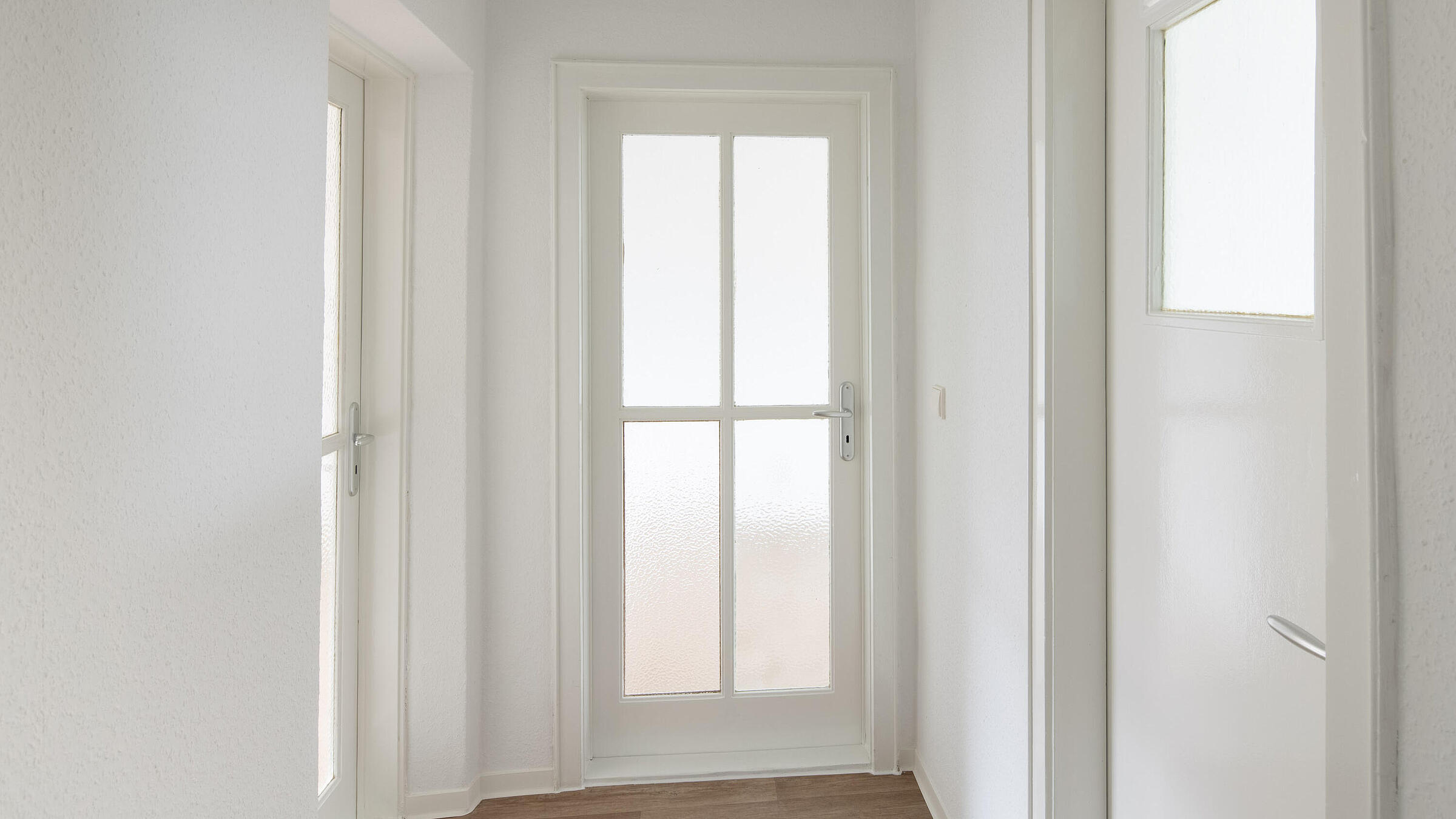 Das Bild zeigt eine modernisierte Wohnung: Ein Flur mit PVC-Belag in Holzoptik, mit einer Tür mit gerostetem Glaseinsatz geradezu, sowie jeweils einer Tür mit Glaseinsatz links abgehend und einer weiteren Tür mit Glasoberlicht rechts abgehend. 