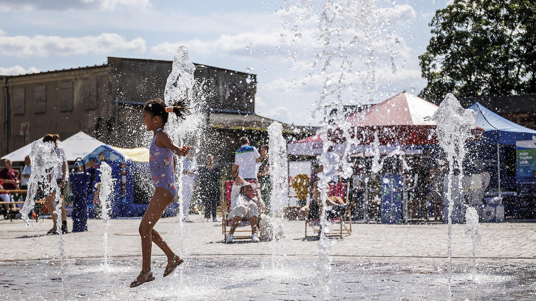 Die Außenaufnahme zeigt den Brunnen auf dem Stadtplatz in Berlin Hellersdorf. Ein Mädchen im Badezug spielt inmitten der kleinen Fontänen mit dem Wasser. Im Hintergrund zu sehen sind vereinzelte Markt- bzw- Informationsstände.