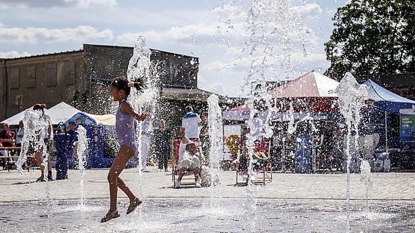Die Außenaufnahme zeigt den Brunnen auf dem Stadtplatz in Berlin Hellersdorf. Ein Mädchen im Badezug spielt inmitten der kleinen Fontänen mit dem Wasser. Im Hintergrund zu sehen sind vereinzelte Markt- bzw- Informationsstände.