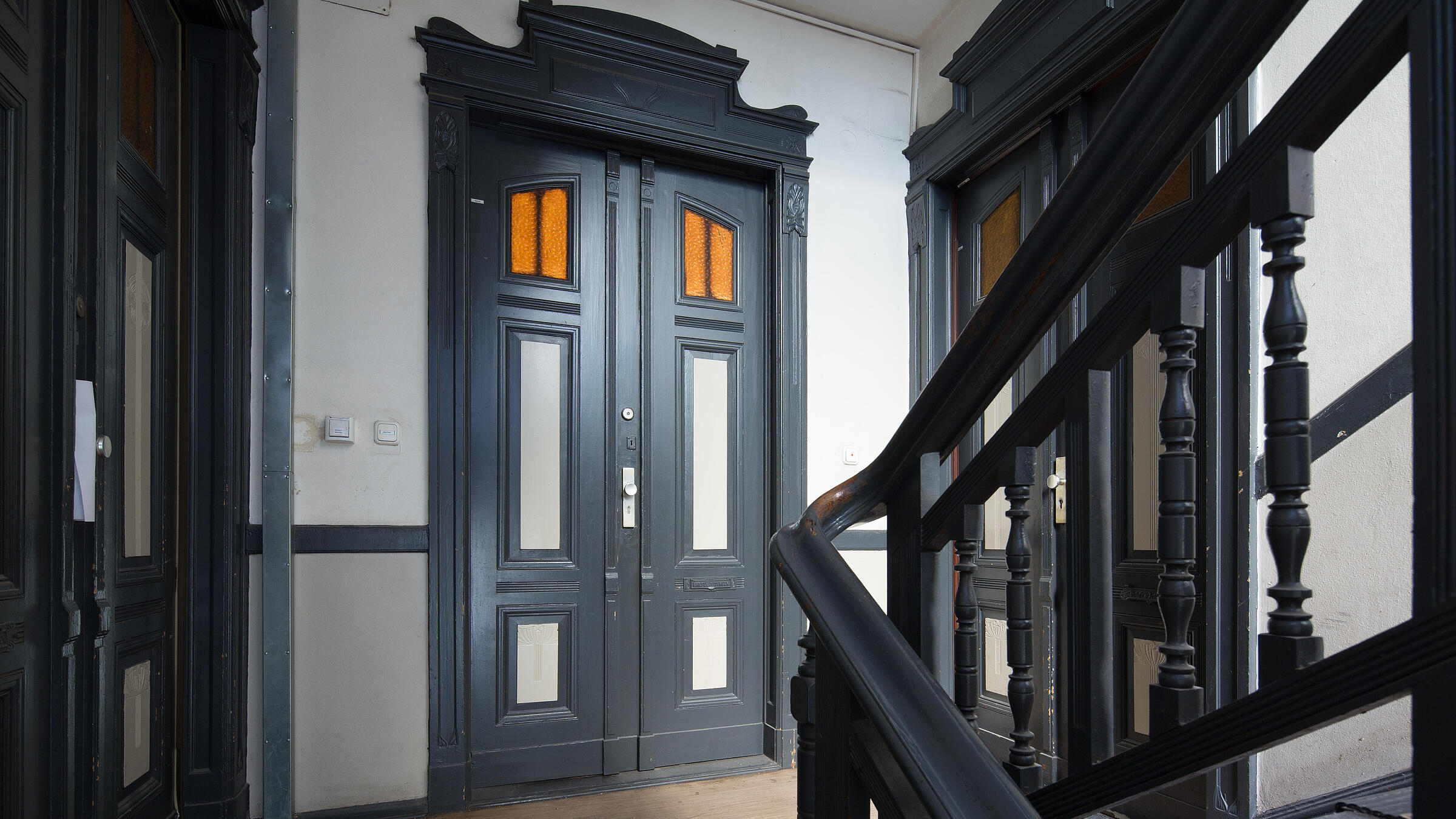 Innenaufnahme zeigt einen Treppenaufgang in der Luxemburger Straße von links die Treppe aufsteigend: Drei Wohnungen mit historisch verzierten Holzeingangstüren, die mittlere als Doppeltür öffnend.