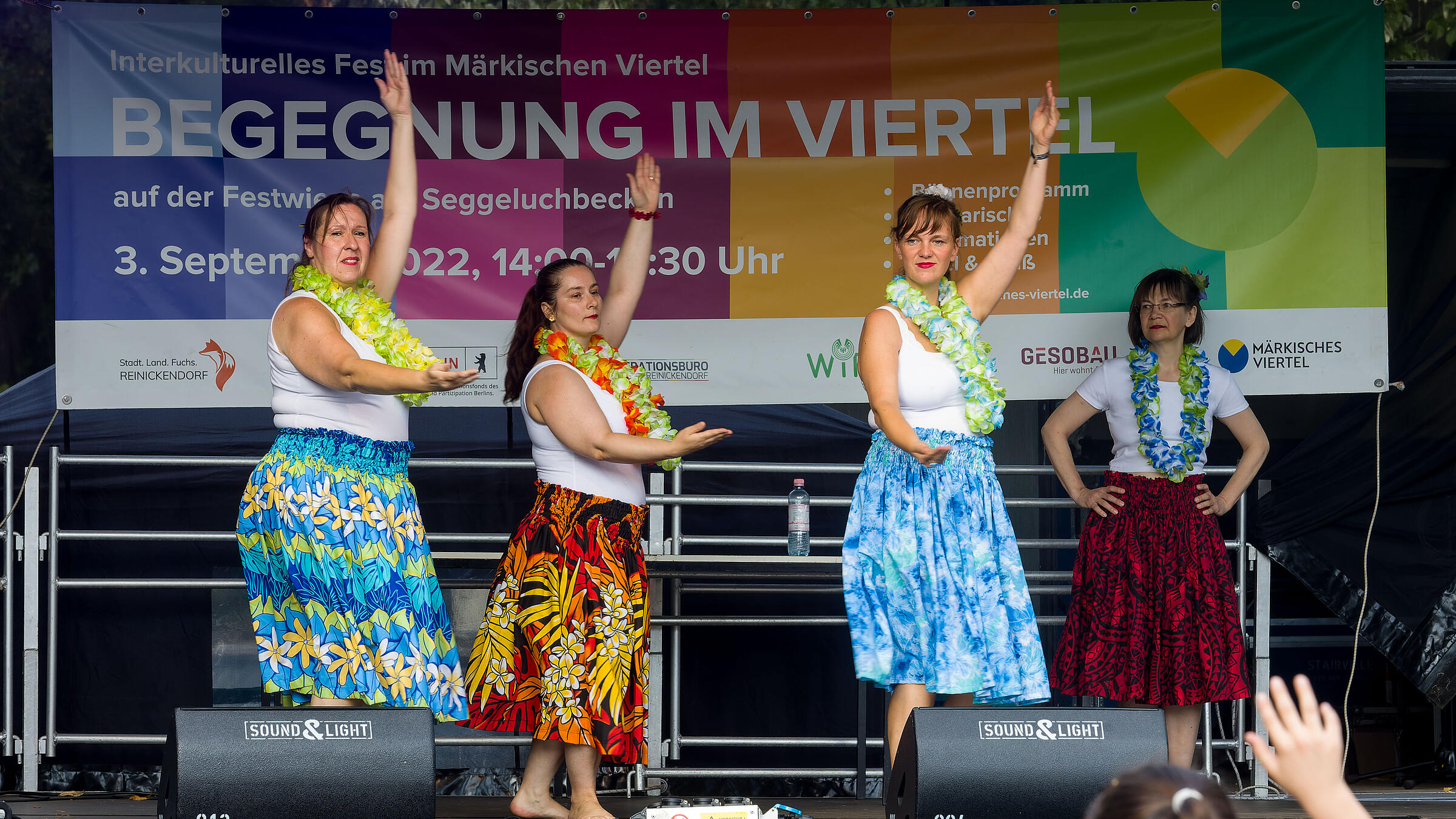 Außenaufnahme zeigt vier Tänzerinnen mit blumigen, langen Röcken und Blumengirlanden auf einer Bühne. 